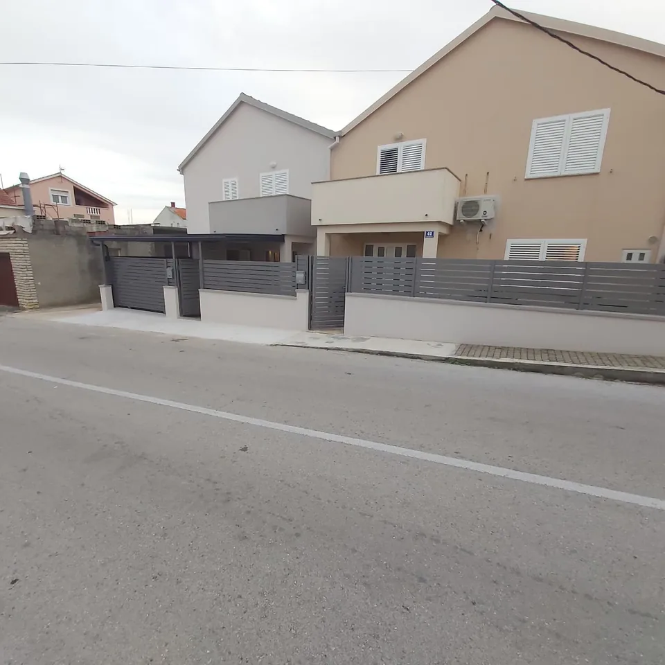 Dvojna kuća sa sivom lamelnom ogradom i nadstrešnicom