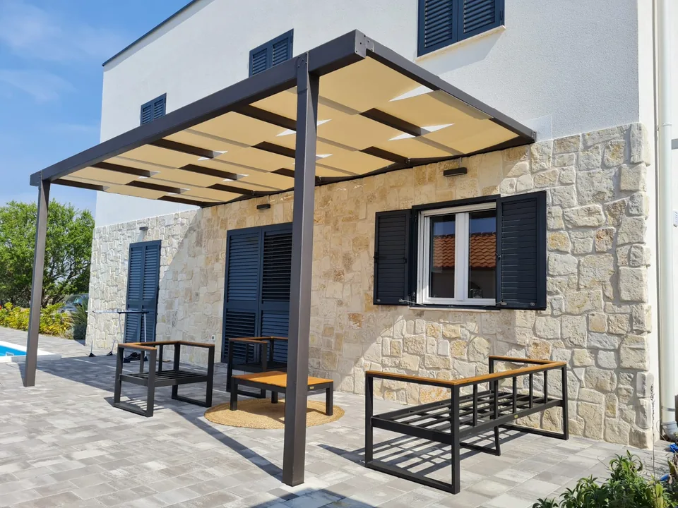 Tamna pergola s platnenom sjenom uz kamenu kuću