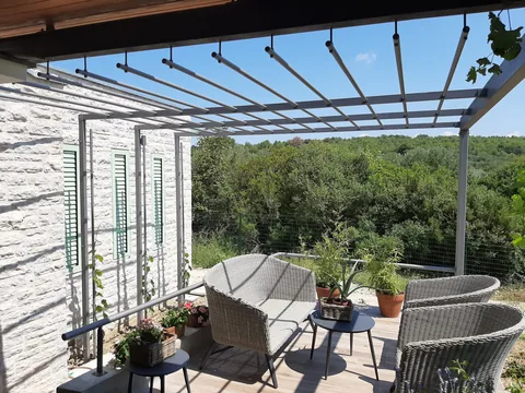 Pergola od sivih kvadratnih profila i okruglih cijevi uz kamenu kuću